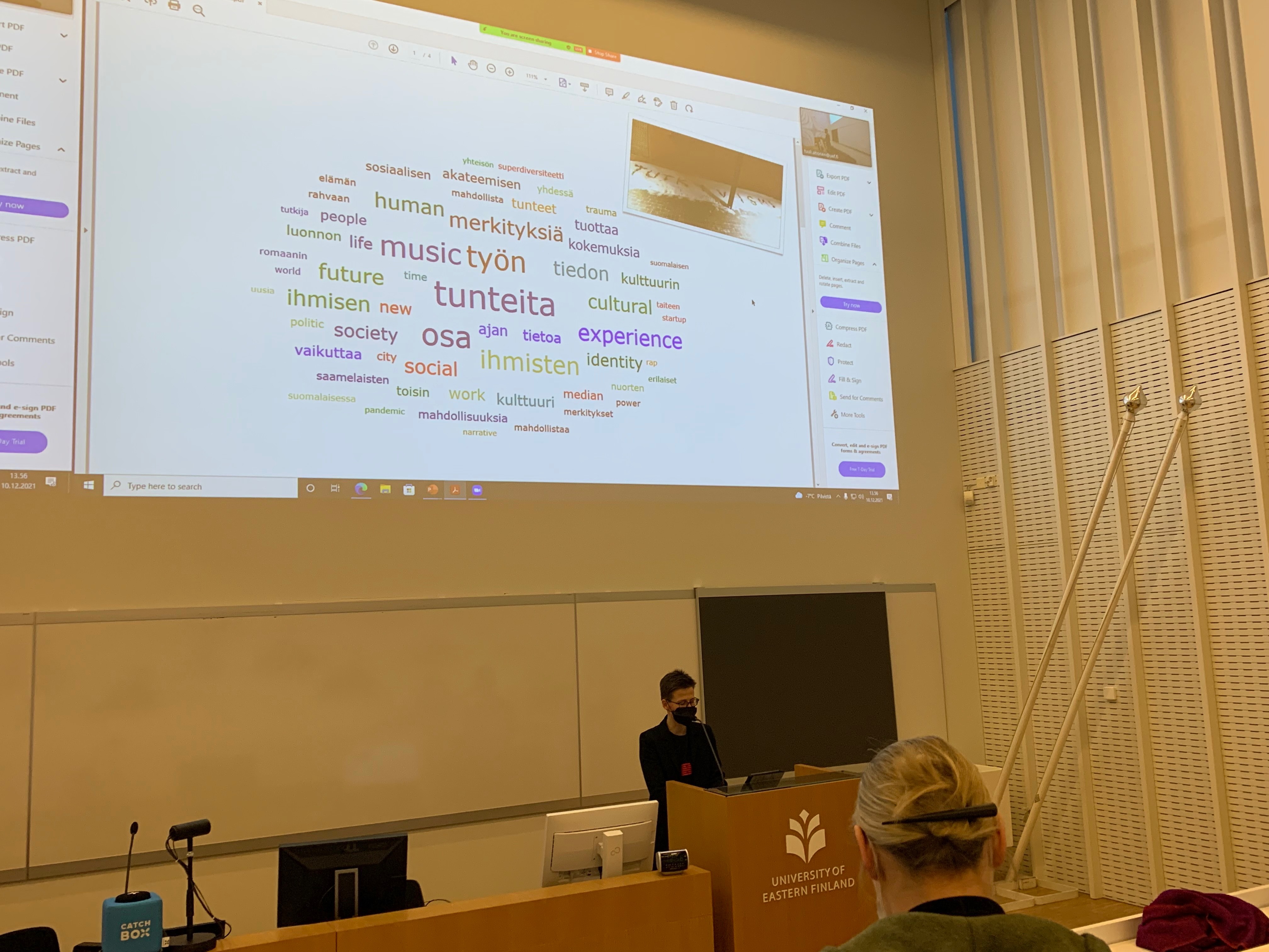 Kuvassa on käynnissä konferenssi, ja ison salin esiintymislavalla seisoo puhuja taustallaan iso valkokangas, jolle on heijastettu sanapilvi. Näytöllä on myös valokuva, jossa lukee ‘tutkivismi’.